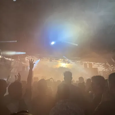 A smoky indoor venue with colorful stage lights and a crowd dancing with hands raised
