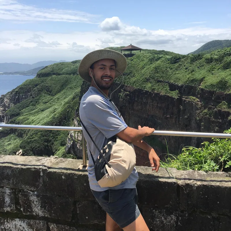 A smiling man in a sun hat leaning on a stone wall overlooking dramatic coastal cliffs and green hills