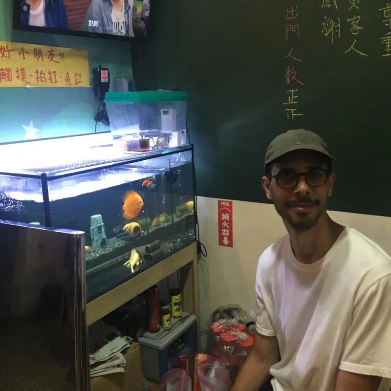 A man in a cap and glasses smiling next to a fish tank with orange discus fish