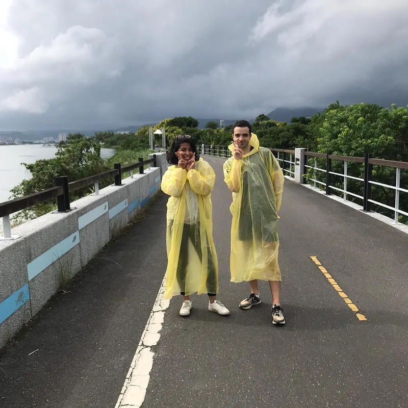 Two people in matching yellow plastic ponchos posing on a riverside path under stormy skies