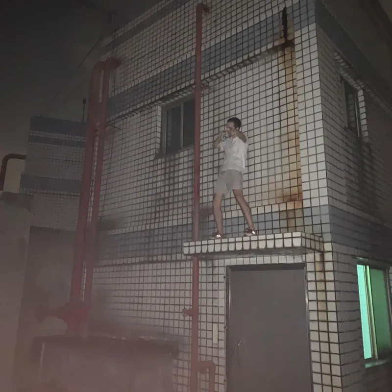 A person standing on a ledge of a tiled building at night, holding a drink