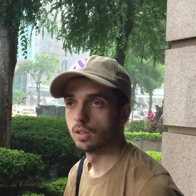 A man in a cap looking sideways in a rainy urban park with trees and hedges behind him