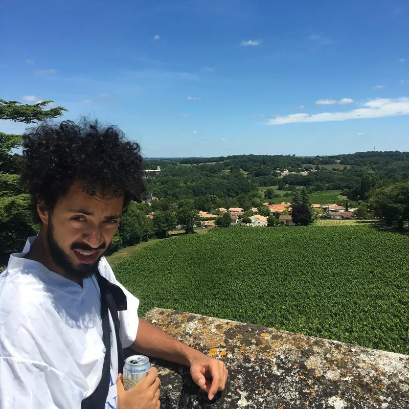 A man with curly hair holding a can, smiling over a stone wall with a sweeping view of vineyards and a village below