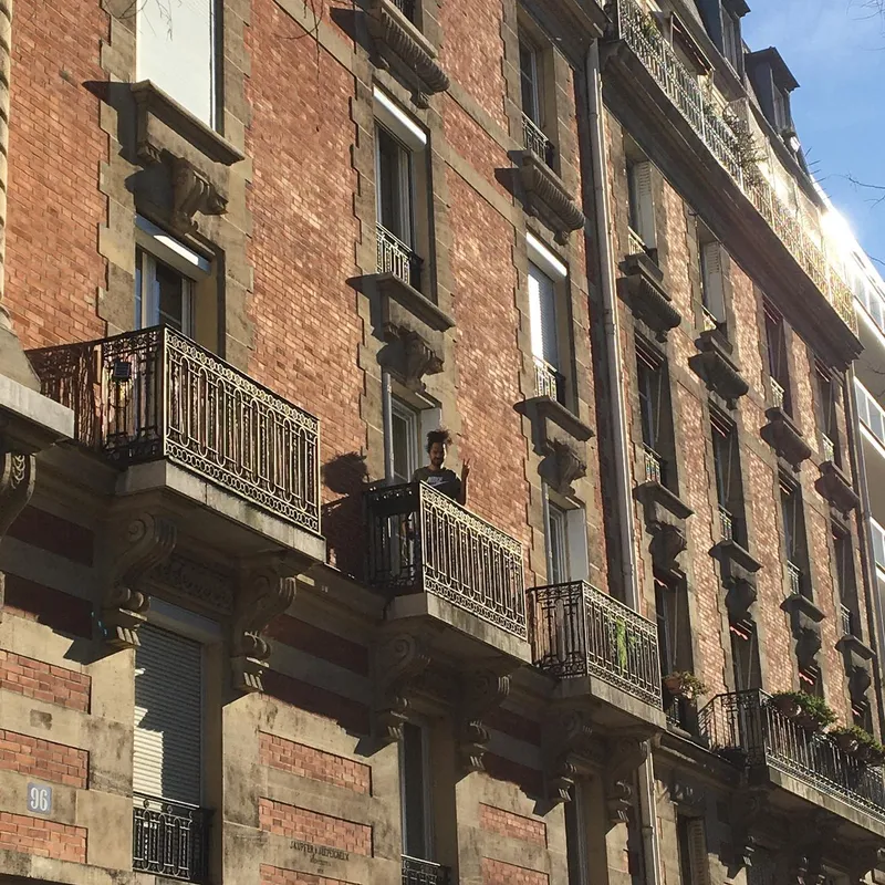 A person standing on a balcony of a red brick Haussmann-style building