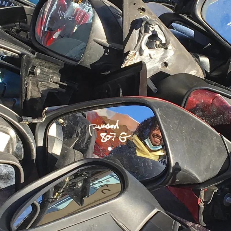 A smiling person reflected in one of many stacked car side mirrors at a market