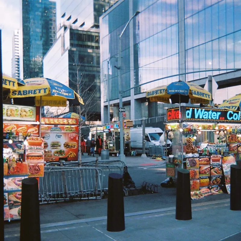 Sabrett hot dog vendor carts lined up on a Manhattan sidewalk with glass skyscrapers reflected behind them