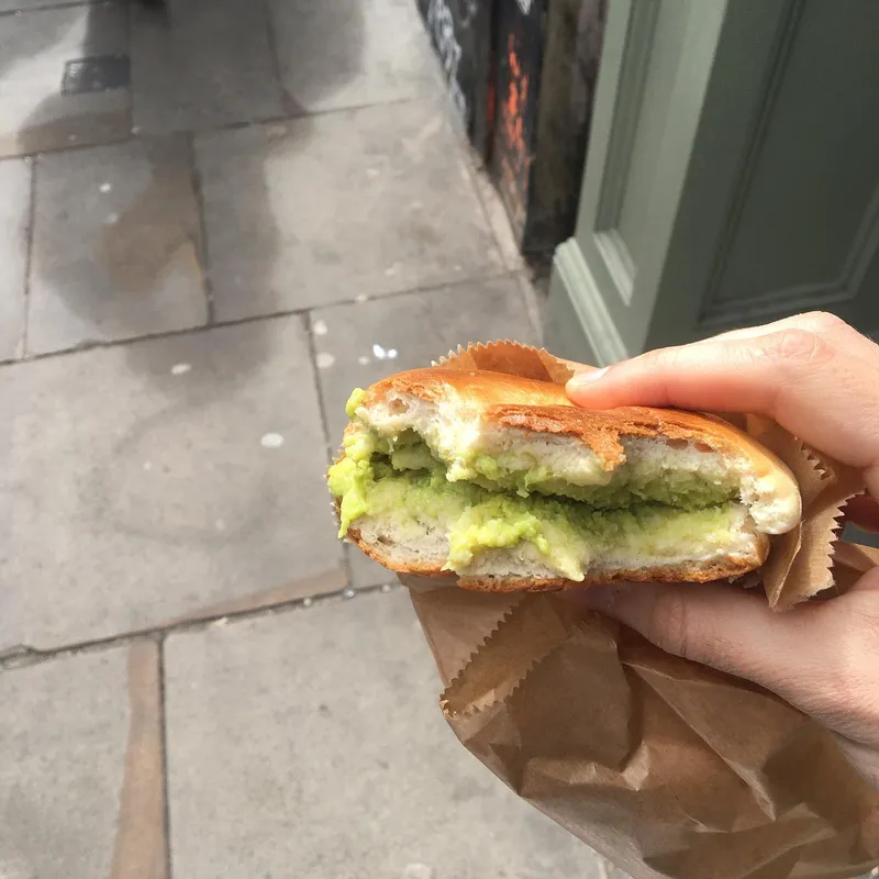 A hand holding an avocado bagel over a pavement