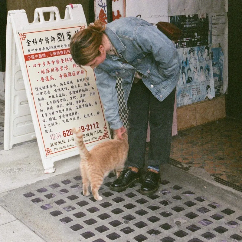 A woman in a denim jacket bending down to pet an orange cat on a Chinatown sidewalk