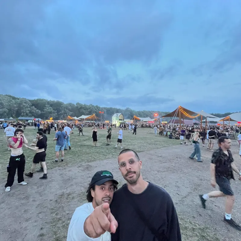 Two friends posing at a festival grounds at dusk with lit tents behind them