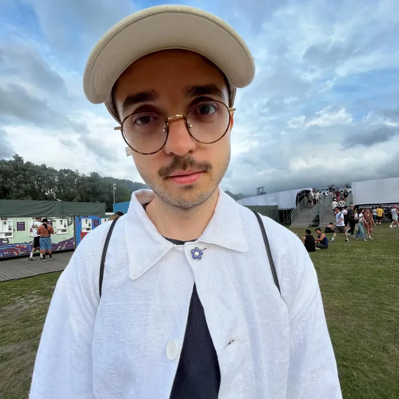A portrait of a young man in a cap and round glasses at a festival
