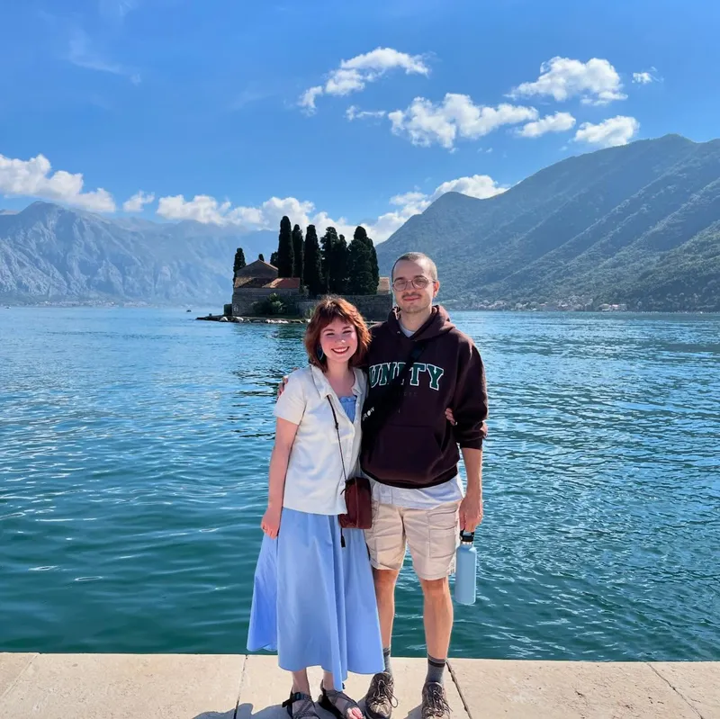 Two people posing on a waterfront with a small island church and mountains behind them