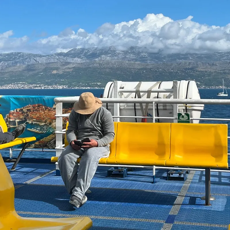 A passenger in a bucket hat sitting on a ferry deck with mountains and sea behind them