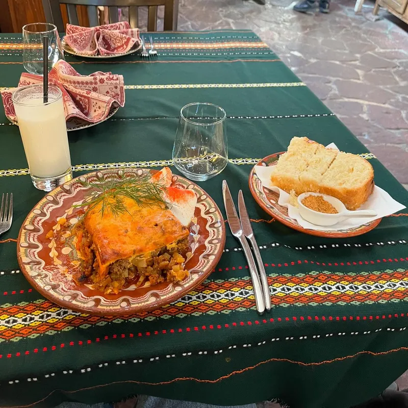 A plate of moussaka with bread and a lemonade on an embroidered tablecloth