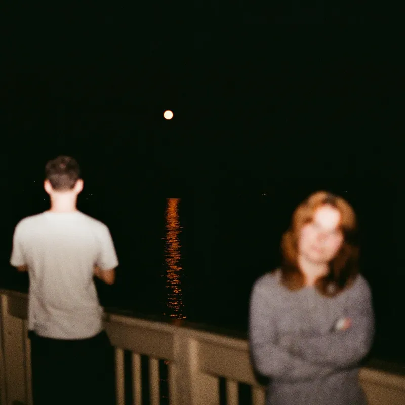 Two figures on a bridge at night with a full moon reflecting on the water